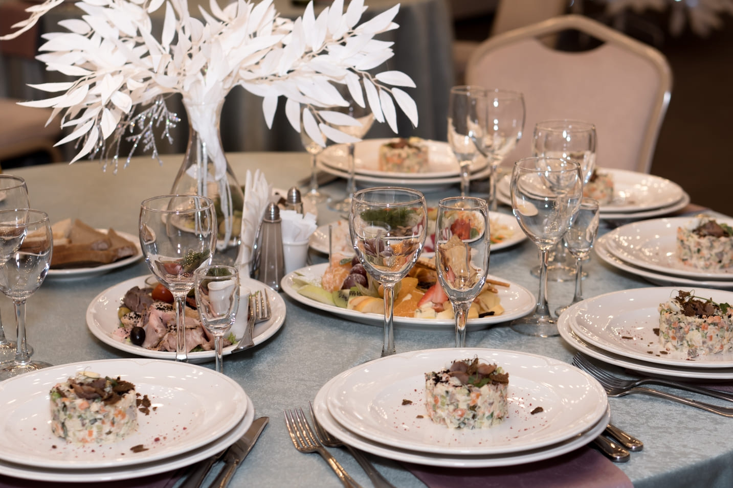 Uma elegante mesa de jantar decorada para uma festa, com pratos de salada servidos em pratos brancos, taças de vinho dispostas ao redor, e um centro de mesa com um arranjo de folhas brancas, criando um ambiente sofisticado e festivo.