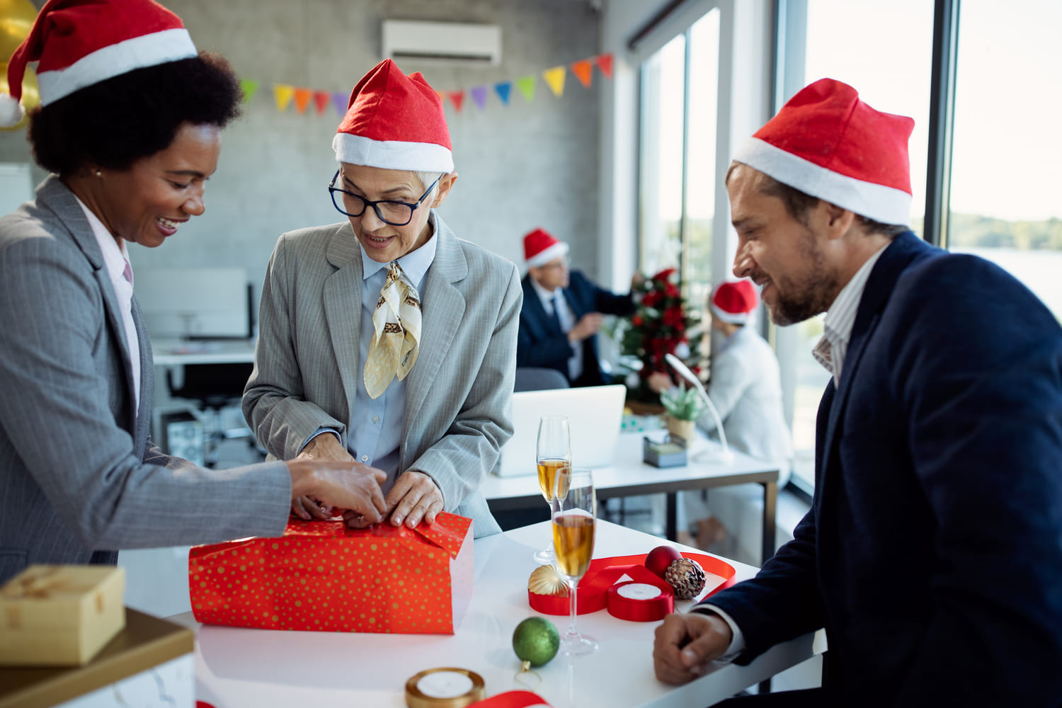 Três pessoas em um ambiente de escritório, usando chapéus de Papai Noel, trocando presentes em uma celebração de Natal. Uma mulher sorri enquanto ajuda a abrir um presente embalado em papel vermelho, enquanto os outros observam. No fundo, outras pessoas decoram uma árvore de Natal.