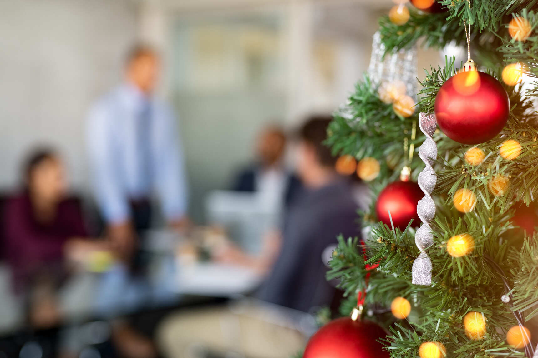 Arvore de natal completamente decorada, ao fundo varias pessoas reunidas em uma reunião com trajes sociais