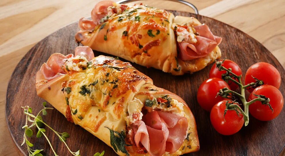 Mini pizza enrolada de mortadela, ao lado um ramo de tomates-cereja, tudo em cima de uma tabua de madeira redonda