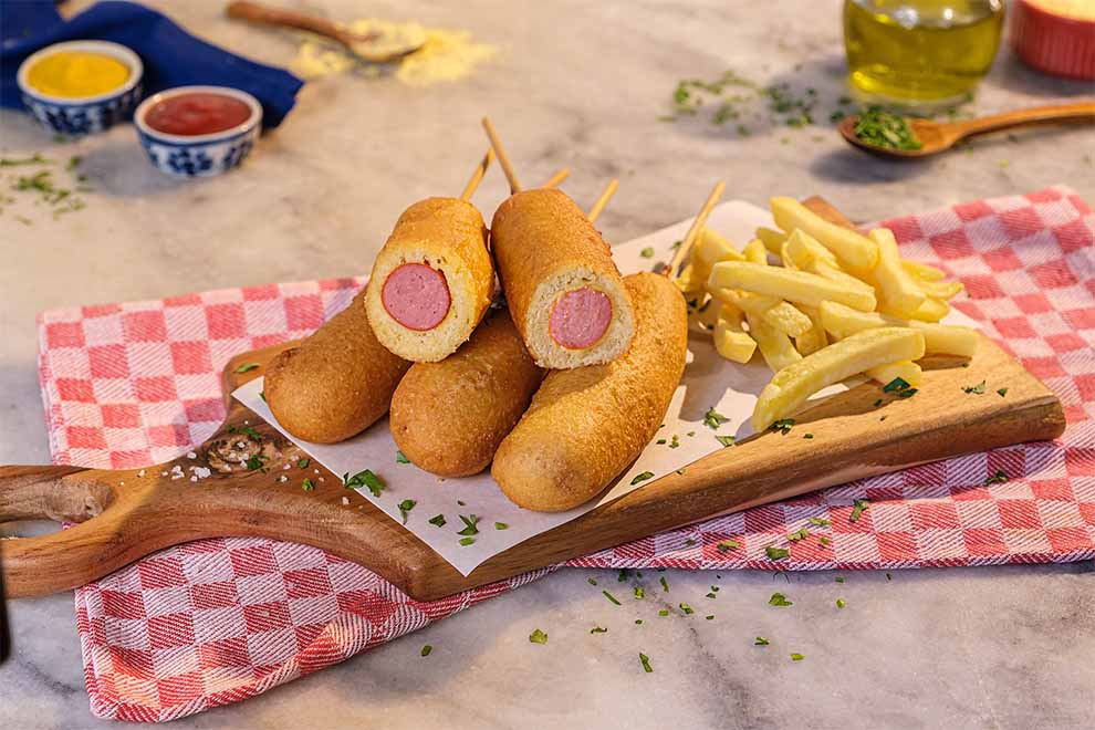 hot dog coreano com fritas ao lado em cima de uma pequena tábua de madeira com um pequeno pano xadrez avermelhado em baixo em uma mesa