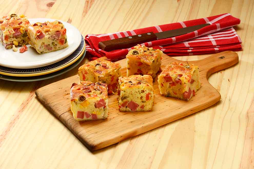 torta de liquidificador com mortadela em cima de uma pequena tábua de madeira e em cima de uma mesa de madeira