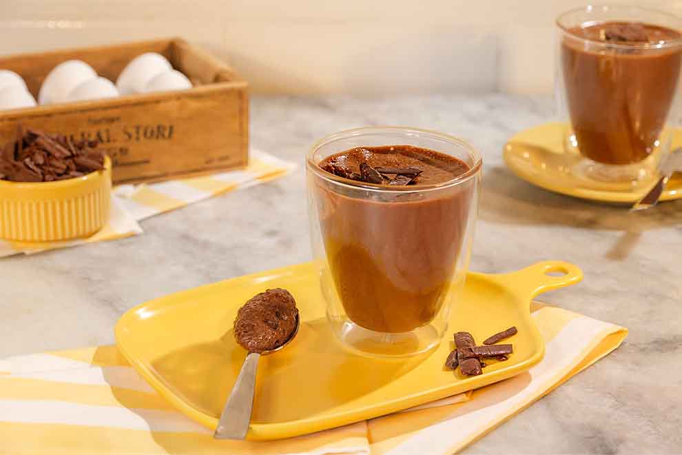 mousse de chocolate dentro de um pequeno copo e uma colher ao lado em cima de um prato retangular amarelo com um pano listrado em amarelo e branco por baixo