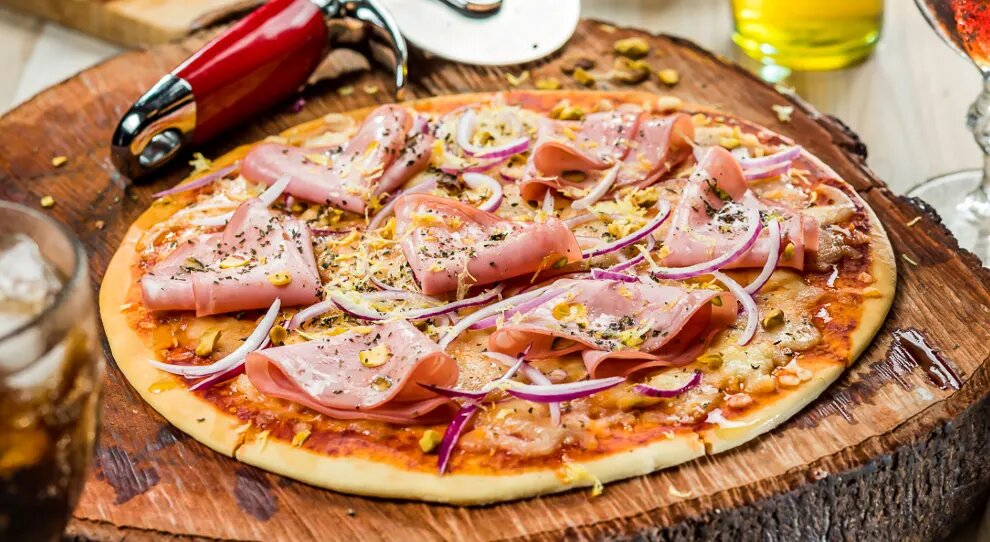 pizza de mortadela e cebola roxa em cima de uma tábua de madeira redonda com um cortador de pizza ao lado em cima de uma mesa de madeira
