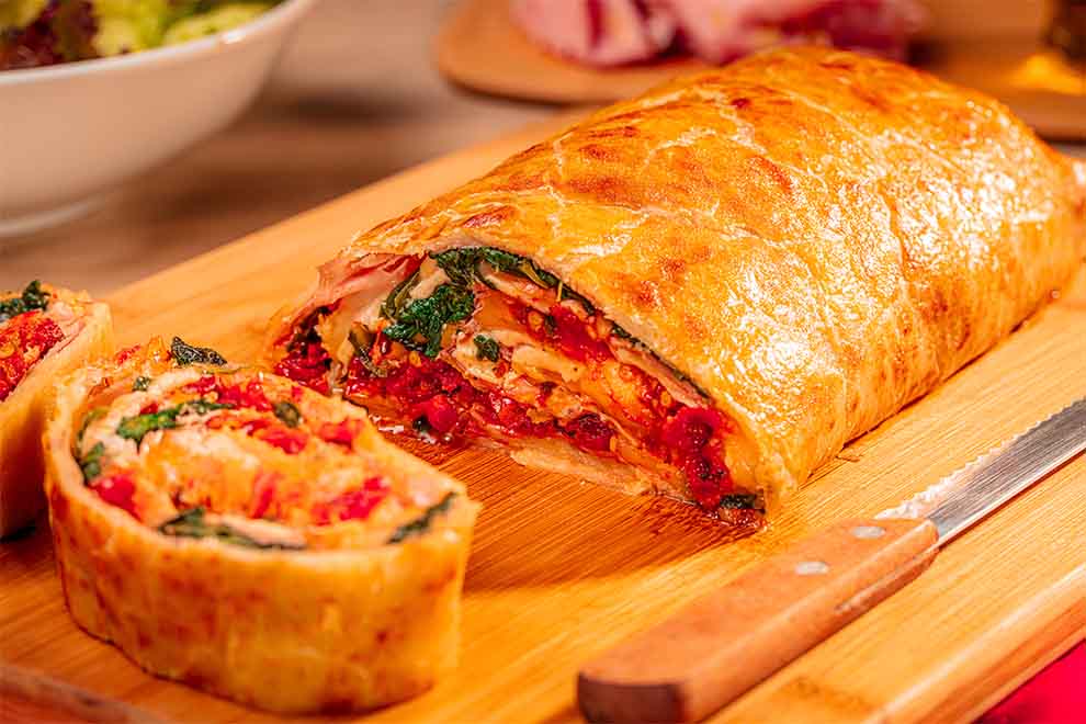 rocambole salgado de mortadela italiana e azeitona em cima de uma tábua de madeira com uma faca ao lado e em cima de um guardanapo vermelho