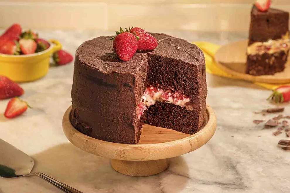 Imagem de um Bolo de chocolate com morango, com uma fatia dele já retirada, em cima de um prato para bolo de madeira, com diversos ingredientes ao redor, sob uma mesa de mármore