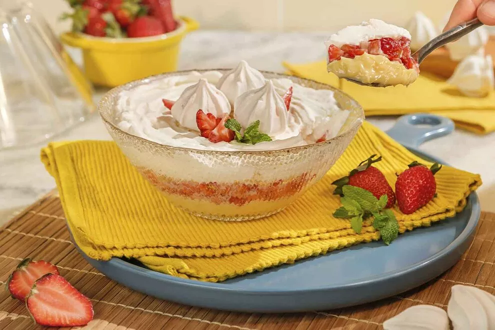 Imagem de um Merengue de morango em uma travessa de vidro de baixo de um pano amarelo dobrado, sob um prato redondo azul, com uma colherada sendo retirada do Merengue, com diversos ingredientes ao redor em cima de uma mesa de mármore