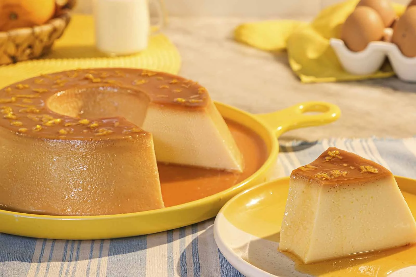 Imagem de um pudim sob uma travessa redonda amarela com uma fatia dele cortada ao lado sob um prato redondo amarelo, ambos em cima de uma mesa de mármore