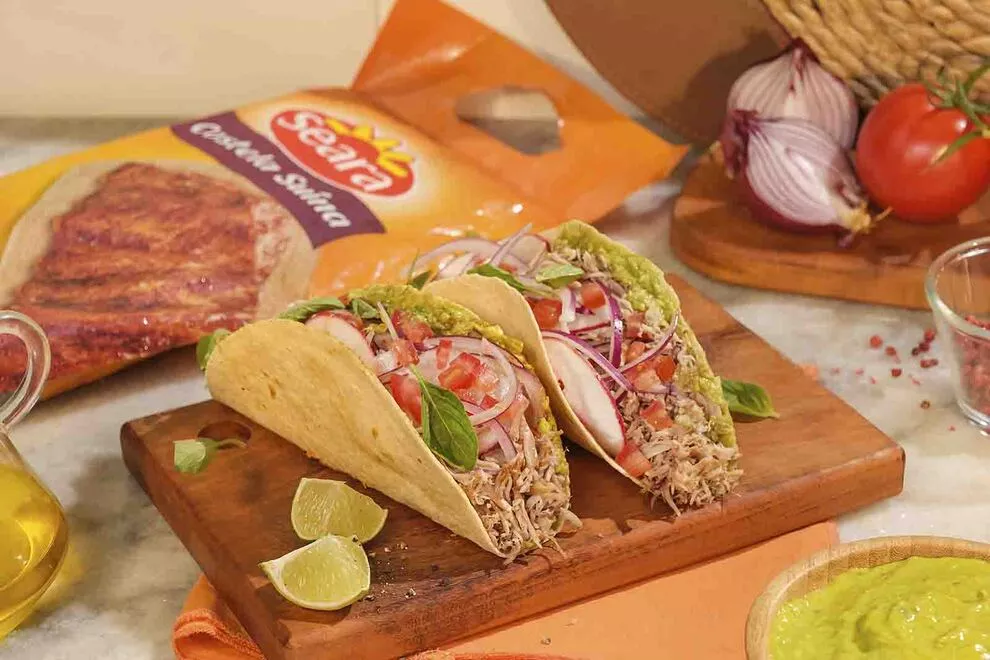 Imagem de duas unidades de Tacos em cima de uma tábua de madeira retangular, com diversos ingredientes ao redor sob uma mesa de mármore