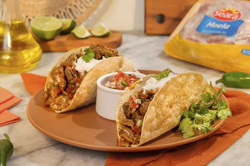 Imagem de duas unidades de Tacos em cima de um prato redondo laranja, com uma combuca de vinagrete no meio, sob uma mesa de mármore com diversos ingredientes ao redor