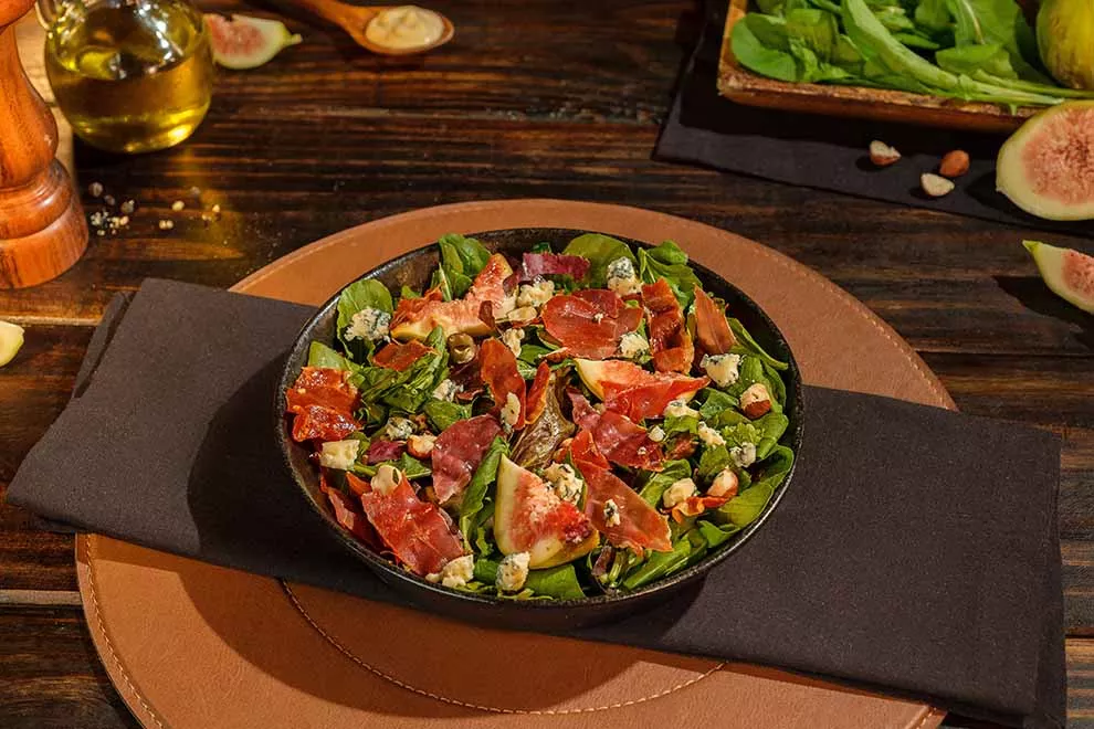 Imagem de um prato escuro com uma Salada verde com presunto cru italiano, sob uma mesa de madeira
