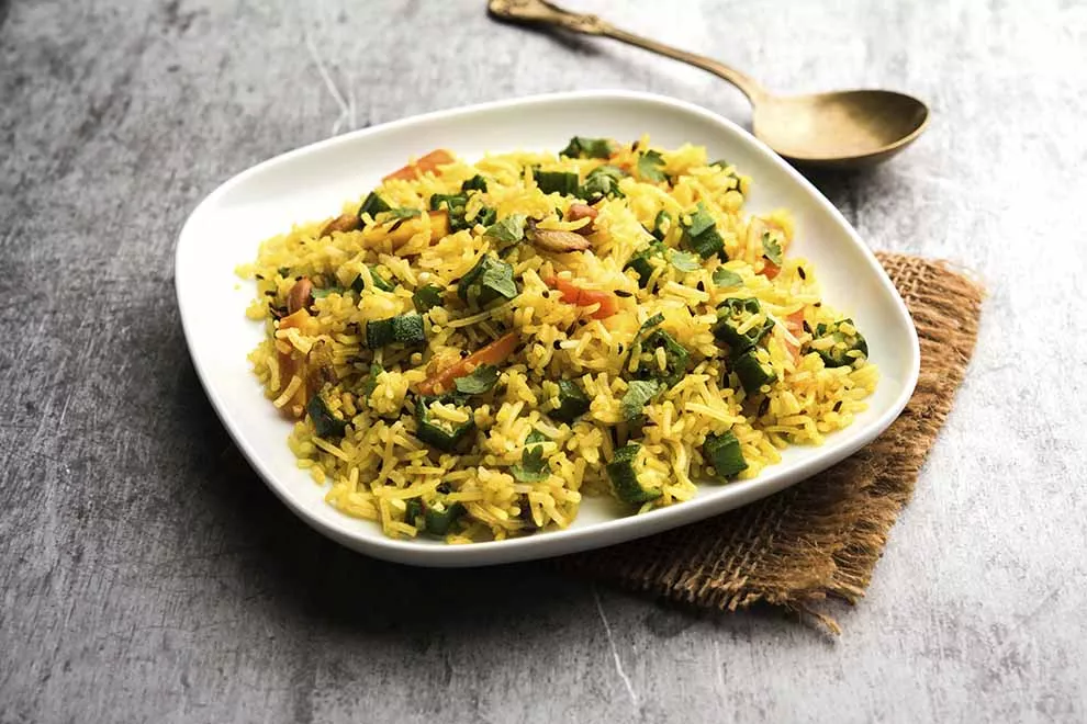 Imagem de um arroz temperado com quiabo em pedaços, sob um prato retangular branco, em cima de um pano marrom dobrado, sob uma mesa cinza