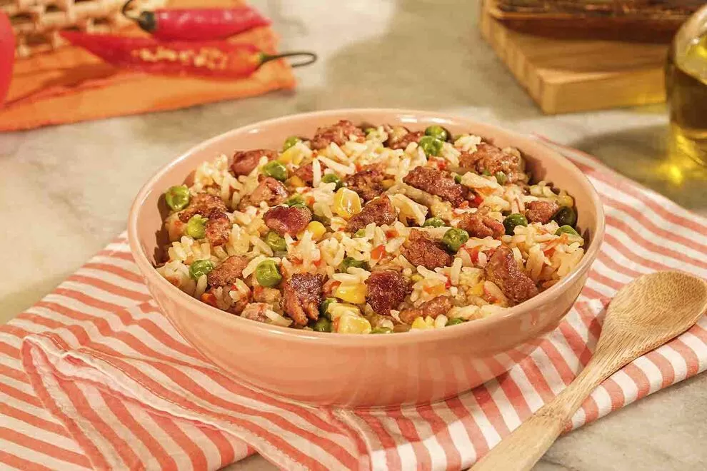 Imagem de um tigela redonda com Arroz com Linguiça Toscana, sob um pano listrado dobrado, em cima de uma mesa de mármore e uma colher de madeira ao lado