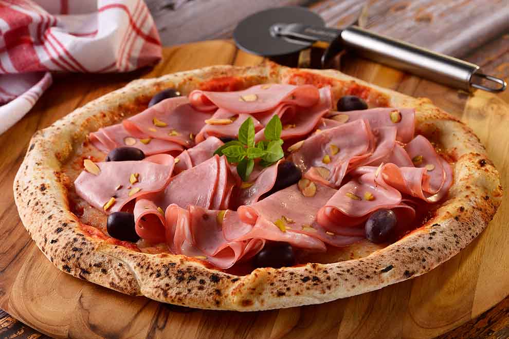 pizza de mortadela e pistache em cima de uma tábua de madeira redonda com um cortador de pizza ao lado e um pano branco com listras vermelhas, em cima de uma mesa de madeira