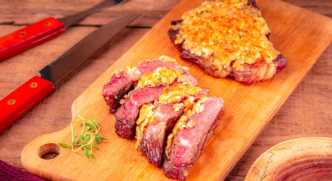 Tábua de madeira marrom com uma porção de bife ancho fatiado por cima. A carne foi assada com uma crosta de alho. Ao lado da tábua, um garfo e uma faca de churrasco.