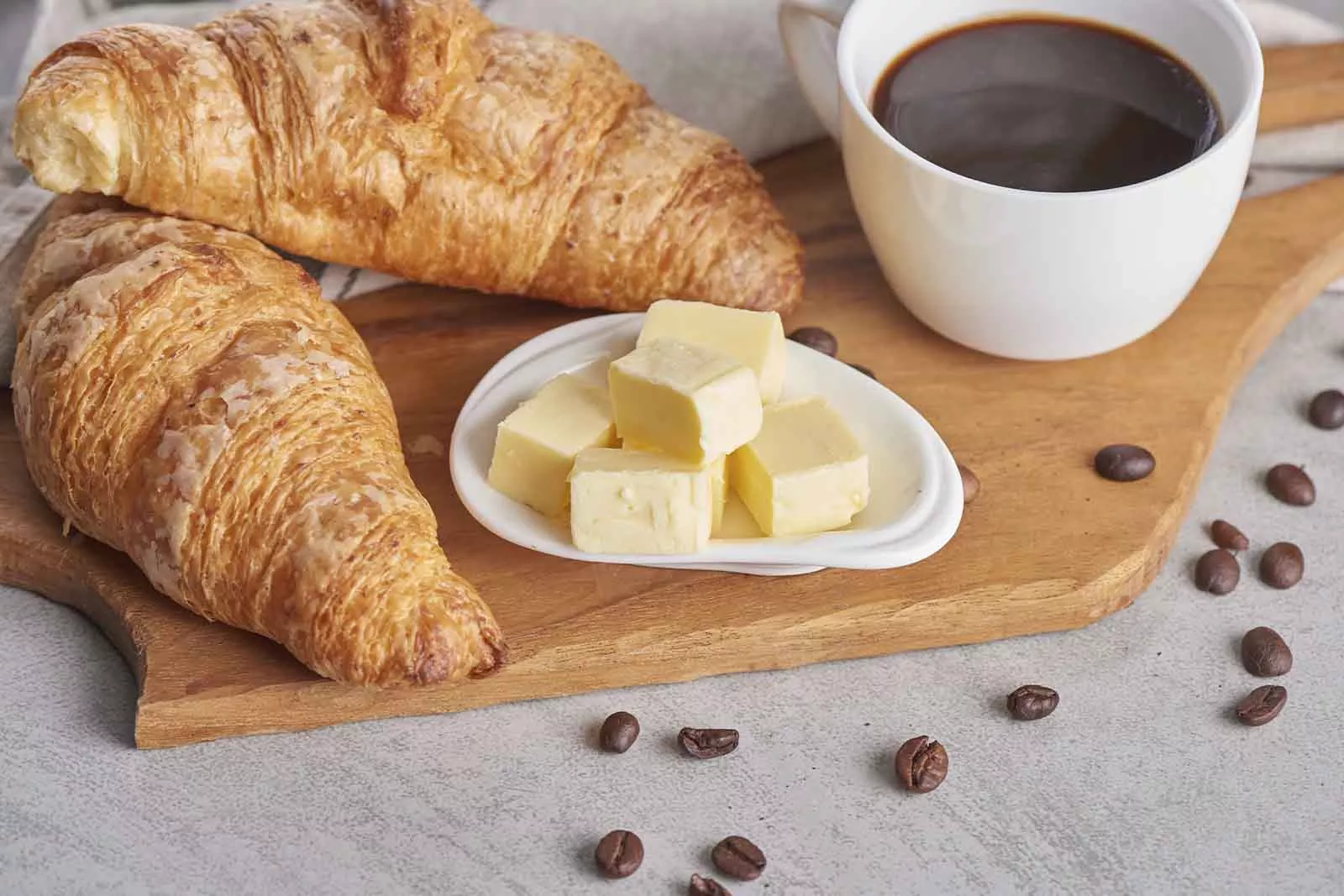 Imagem de duas unidades de Croissant,  com uma xícara de café ao lado e alguns pequenos cubos de manteiga sob um pequeno pires, em cima de uma tábua de madeira, em cima de uma mesa cinza com alguns grãos de café espalhadas pela mesa