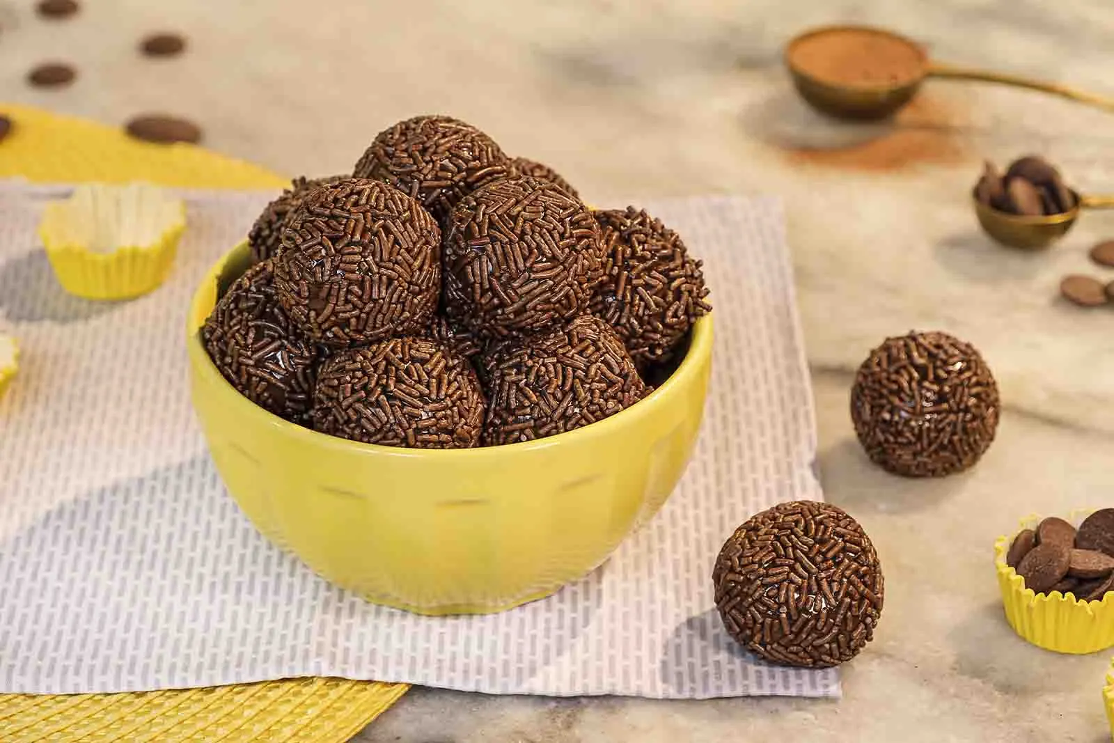 Imagem de diversos brigadeiros tradicionais, em cima de uma tigela amarela, sob uma mesa de mármore