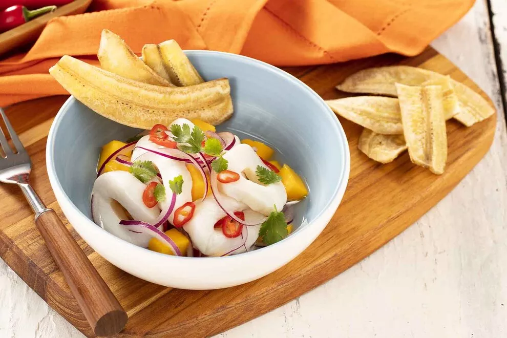 Imagem de um Ceviche de lula com manga, em cima de uma tigela pequena azul, com um garfo ao lado e chips de banana, em cima de uma tábua de madeira retangular, sob uma mesa de madeira branca