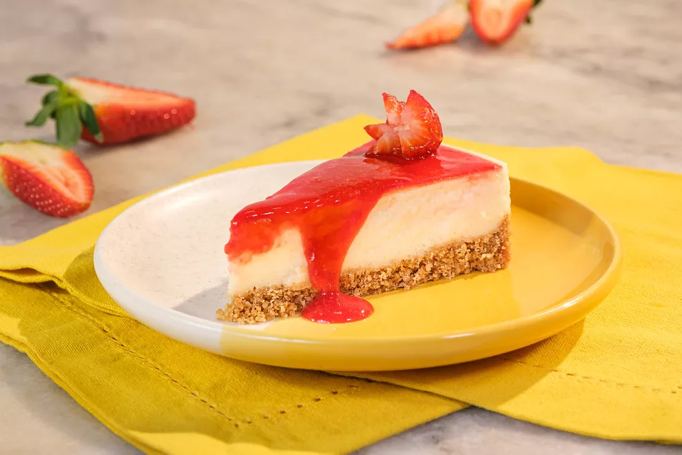 Imagem de uma fatia de Cheesecake de morango em cima de um prato redondo amarelo, sob uma toalha amarela dobrada, todos sob uma mesa de mármore