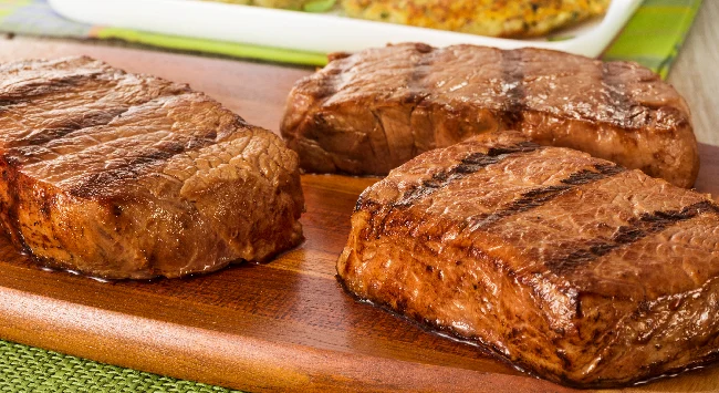 Três pedaços de bife de contrafilé grelhados e dispostos em uma tábua de madeira. Ao fundo, uma travessa com abobrinhas recheadas.