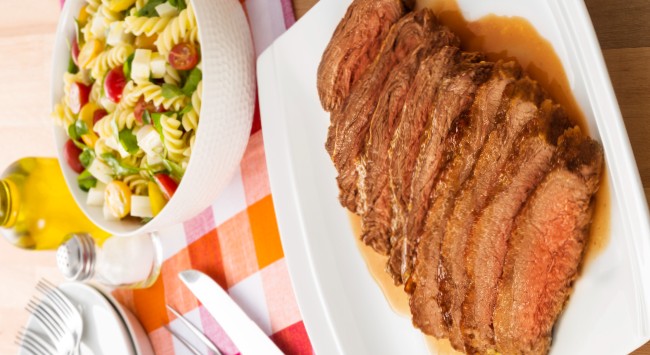 Fatias de fraldinha servidas em um prato retangular, branco e raso. Atrás, uma tigela branca simples com salada de macarrão e atrás delas um saleiro e um frasco de azeite. Do lado esquerdo disso, dois pratos, um em cima do outro com três garfos em cima. Mais à frente dos pratos, entre eles e o prato de fraldinha, um par de garfo e faca. Os elementos estão sobre uma mesa forrada com pano vermelho, laranja e branco quadriculado.