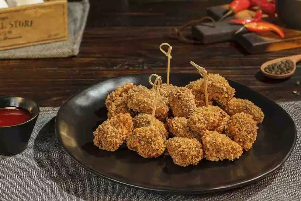 Imagem de diversos corações de galinha empanados, em cima de um prato redondo escuro, com diversos ingredientes ao redor e uma combuca de molho ao redor, sob uma mesa de madeira 