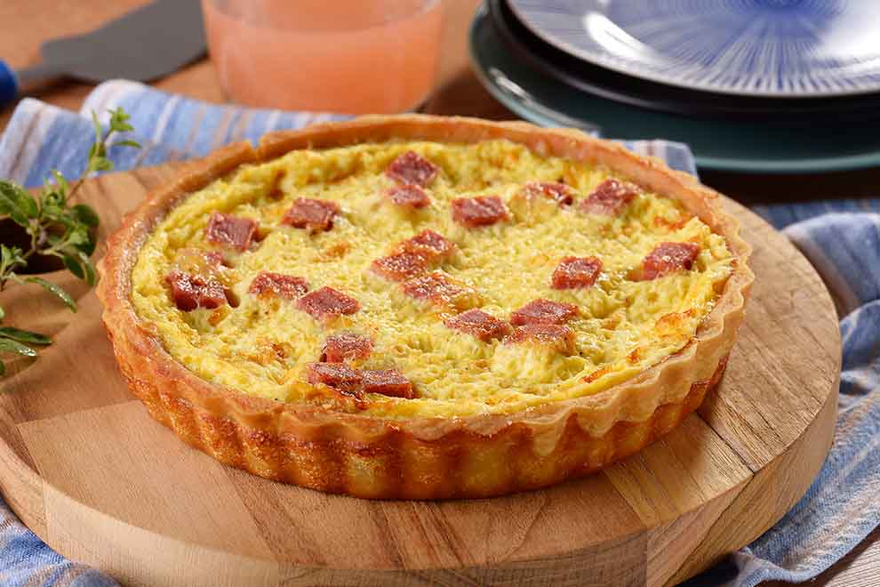 quiche de mortadela e queijo fresco em cima de uma tábua redonda de madeira com um pano azul claro embaixo e em cima de uma mesa de madeira