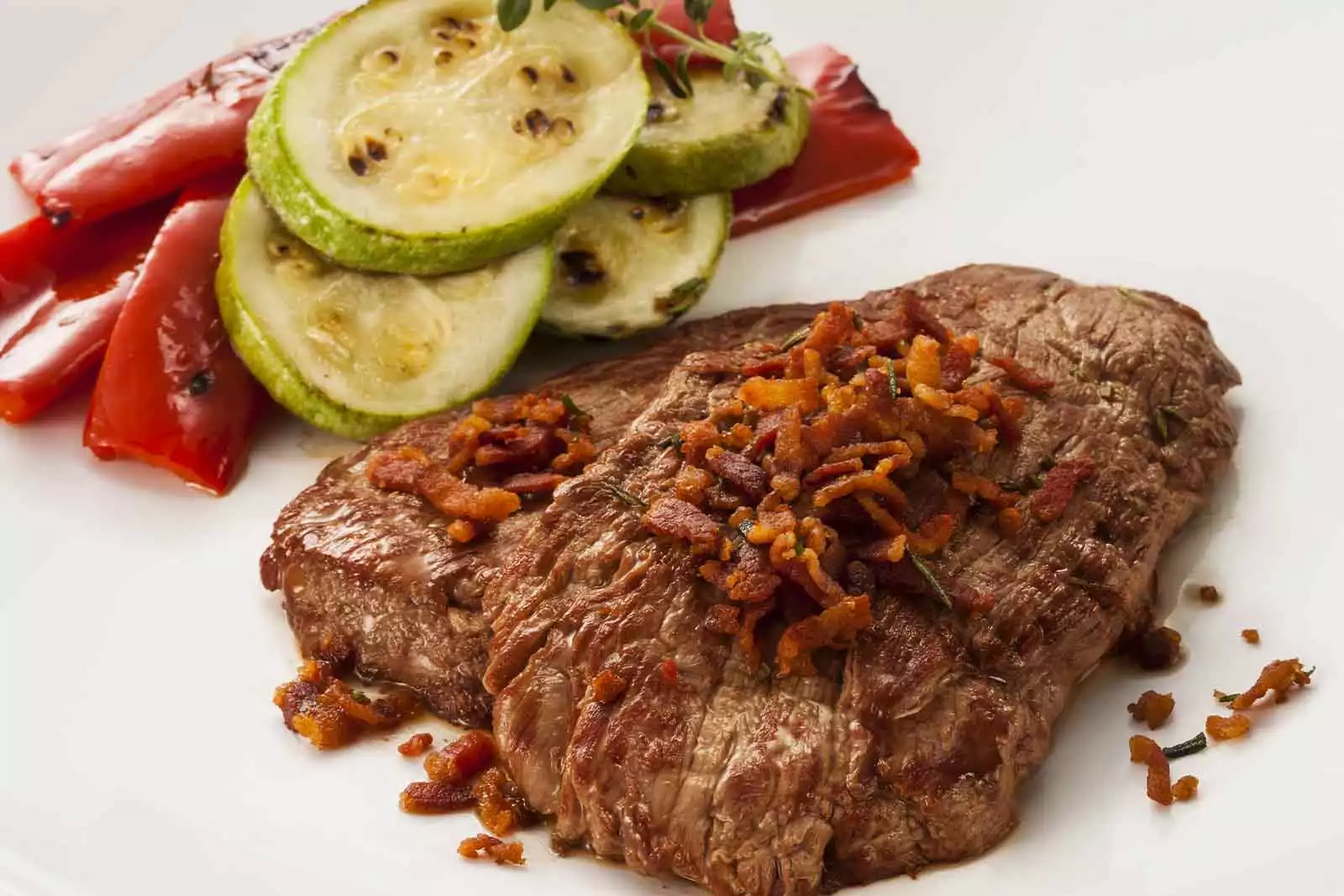 Imagem de um Escalope de filé mignon com bacon crocante, com alguns legumes assados ao redor, sob uma prato redondo branco