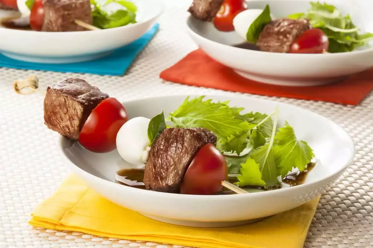 Imagem de um espetinho caprese em cima de um pequeno prato redondo branco, em cima de um pano amarelo dobrado, sob uma mesa branca com outros espetinhos caprese ao fundo