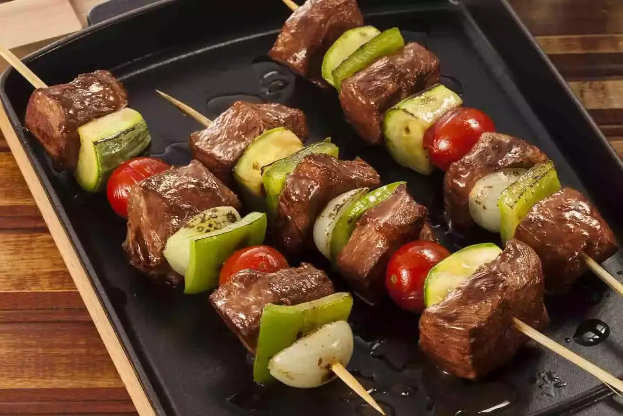 Imagem de três unidades de espetinho de paleta, sob uma travessa de metal retangular, em cima de um suporte de madeira, sob uma mesa de madeira