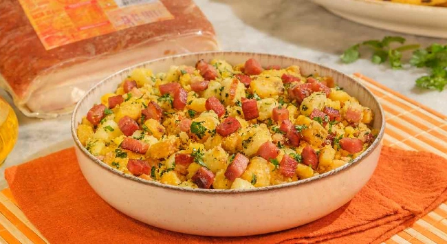 Tigela de farofa de cuscuz com bacon por cima de um guardanapo laranja, em um jogo americano listrado amarelo e laranja. No fundo da imagem, uma embalagem de bacon mantinha Seara