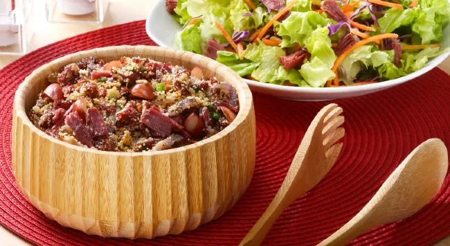 Farofa de Jerked Beef com pinhão, servida em uma tigela de madeira, que contrasta bem com o tecido vermelho que serve como base.