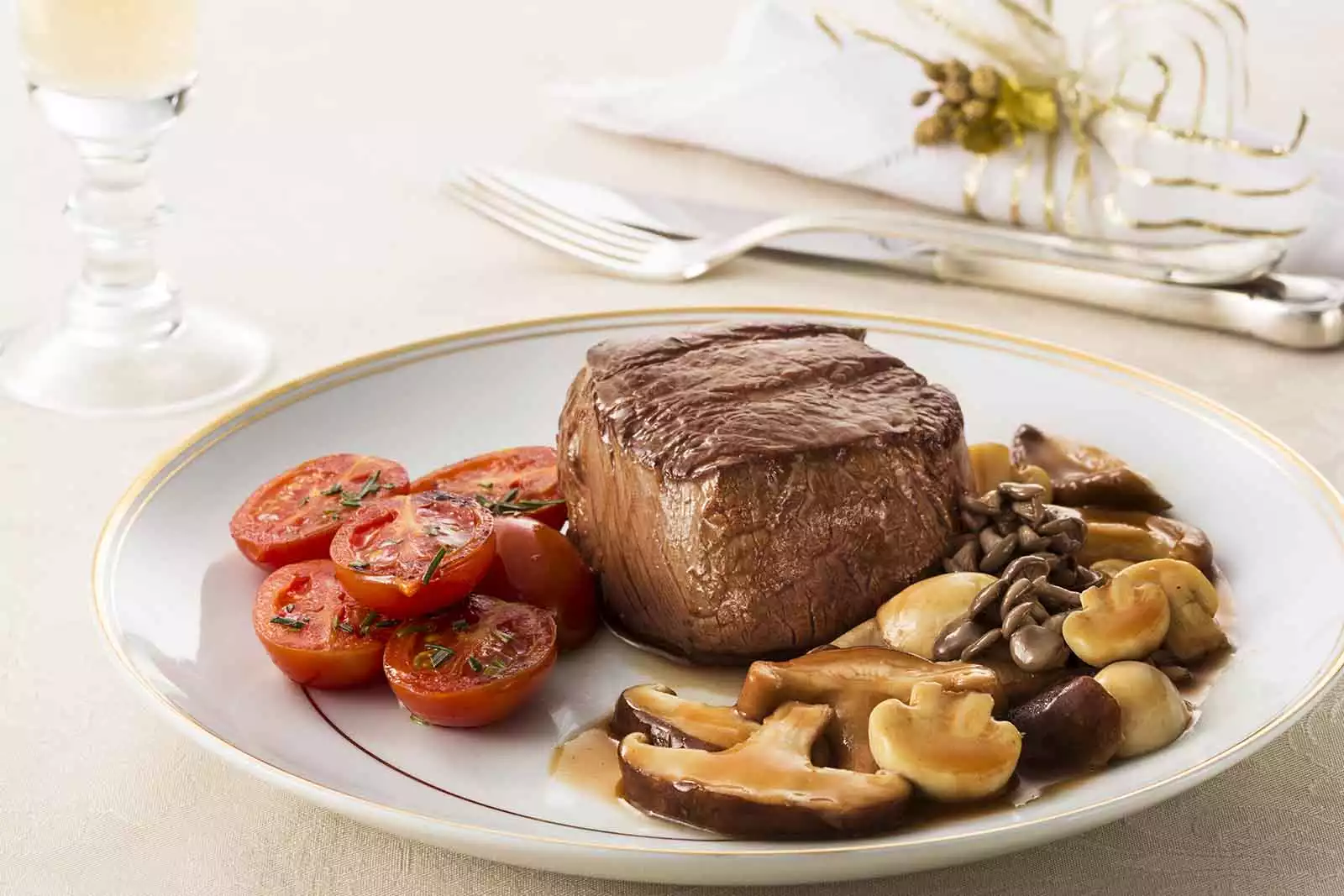 Imagem de uma peça de Filé mignon assada, com diversos cogumelos e tomates ao redor, sob um prato redondo branco, em cima de uma mesa com toalha branca, um par de talheres ao lado, um guardanapo de pano e a base de uma taça de vidro no canto esquerdo da imagem