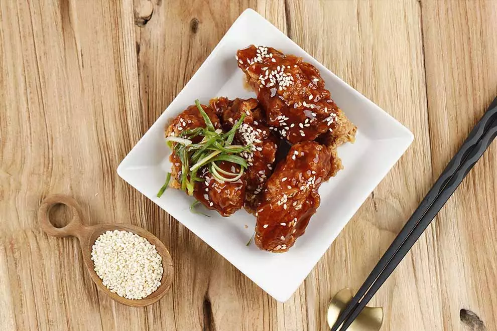 Imagem de um Orange chicken em cima de um prato quadrado branco, com um par de hashis e uma combuca de gergelim ao lado, sob uma mesa de madeira