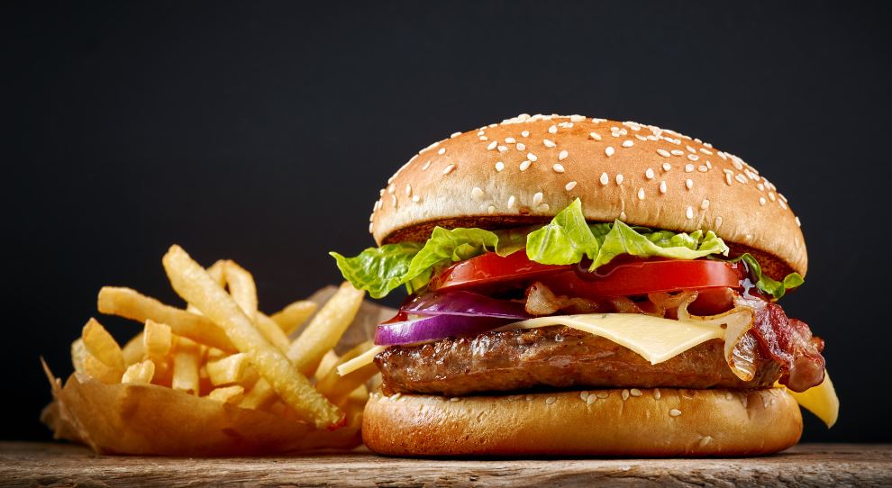 Hambúrguer artesanal motado com alface, tomate, queijo e cbola no pão com gergelim, ao lado de uma porção de batatas fritas em uma tábua de madeira.