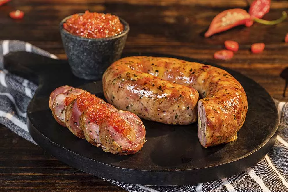 Imagem de uma unidade de linguiça cuibana, com uma parte dela cortada em fatias com molho por cima, sob uma tábua de madeira escura, com uma pequena combuca de molho ao lado, sob um pano dobrado listrado, em cima de uma mesa de madeira