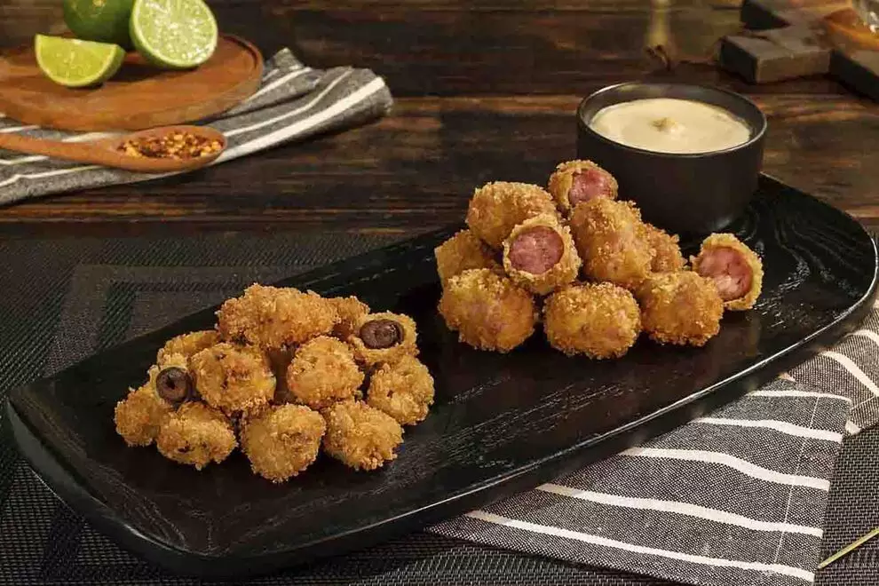 Imagem de diversas Linguiças e Coraçôes de Frango Empanados, sob uma prato retangular escuro, com uma combuca de molho ao lado, sob uma mesa de madeira