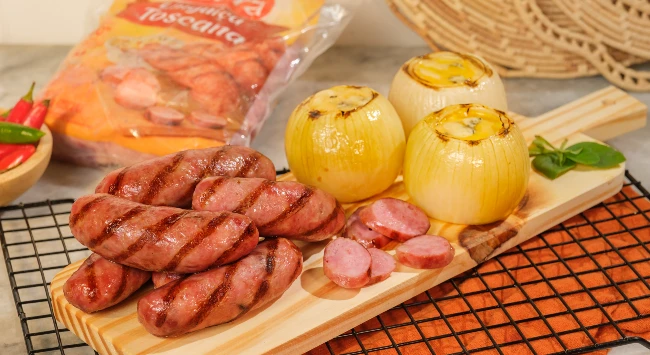 Tábua de madeira com linguiças toscanas grelhadas, com marcas de grelha visíveis, acompanhadas de cebolas inteiras assadas.