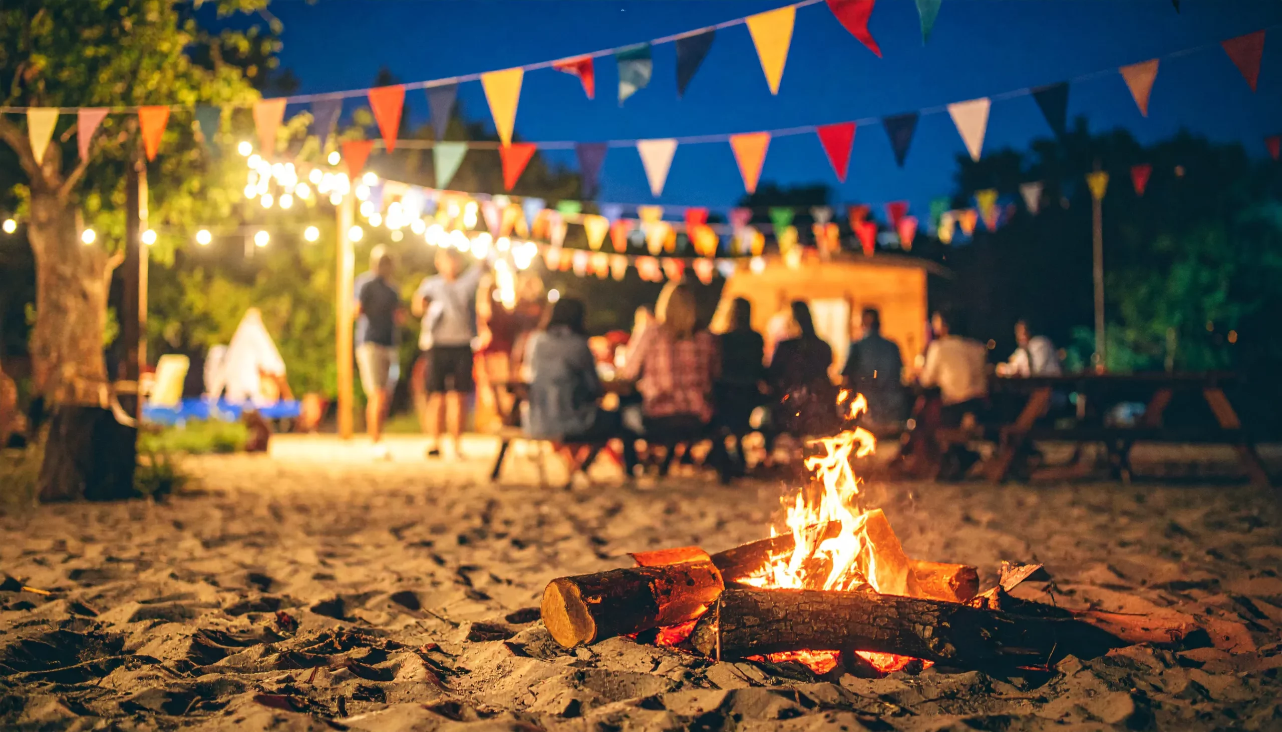 Uma fogueira acesa na areia em primeiro plano, com pessoas ao fundo em mesas sob luzes e bandeirinhas coloridas, compondo a decoração para festa junina.