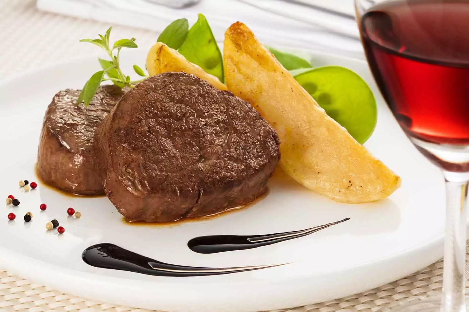 Imagem de duas unidades de Filé mignon assados, com duas unidades de batatas rusticas, folhas de salada, temperos e molho balsâmico ao redor, sob um prato redondo branco, com uma taça de vinho no canto direito da imagem