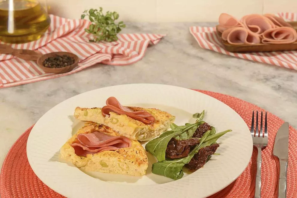 Imagem de um omelete de forno com rúcula com tomate seco ao lado, sob um prato redondo branco sob uma jogo de mesa redondo rosa, em cima de uma mesa de mármore com um par de talheres ao lado