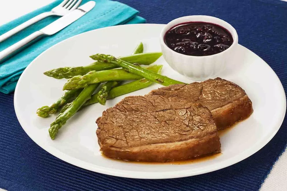 Imagem de duas fatias de picanha assadas com diversos aspargos assados ao lado e uma combuca de molho, sob um prato redondo branco sob uma mesa com pano azul e um pra de talheres ao lado