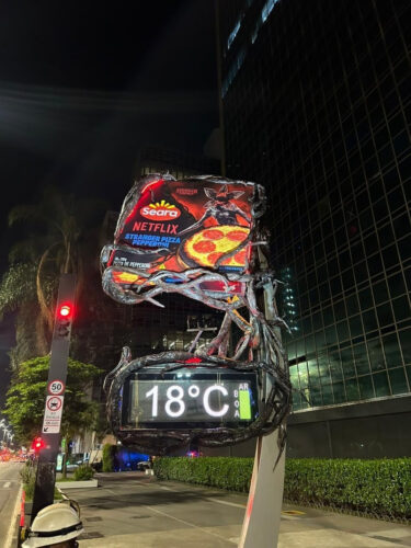 Painel de temperatura coberto por uma instalação inspirada em Stranger Things, com uma embalagem gigante de pizza Seara Stranger Pizza pepperoni no topo, em uma rua à noite.